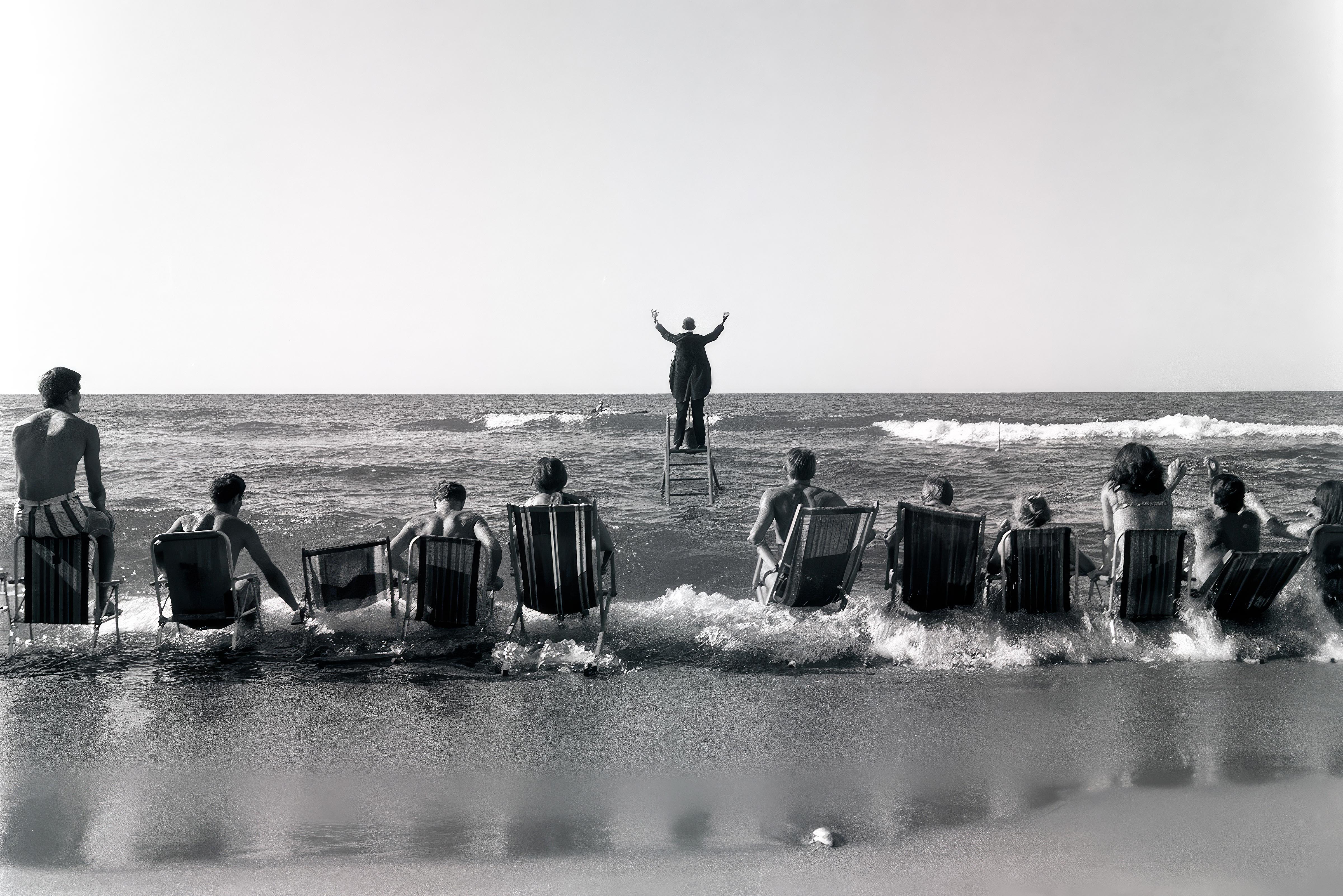 Eine Gruppe von Menschen sitzt in Liegestühlen am Strand, mit Blick auf das Meer. Im Vordergrund stehen die Stühle teilweise im Wasser. Im Hintergrund steht eine Person auf einem Podest oder Stuhl, mit erhobenen Armen, dem Meer zugewandt. Die Szene ist in schwarz-weiß gehalten, das Wasser zeigt leichte Wellen.